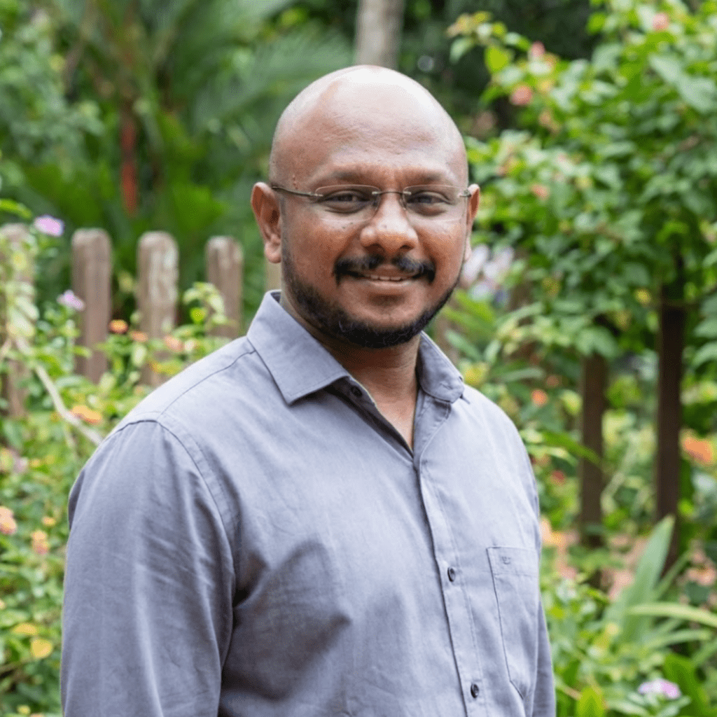 A bald man with glasses and a beard smiling in a garden with a wooden fence.