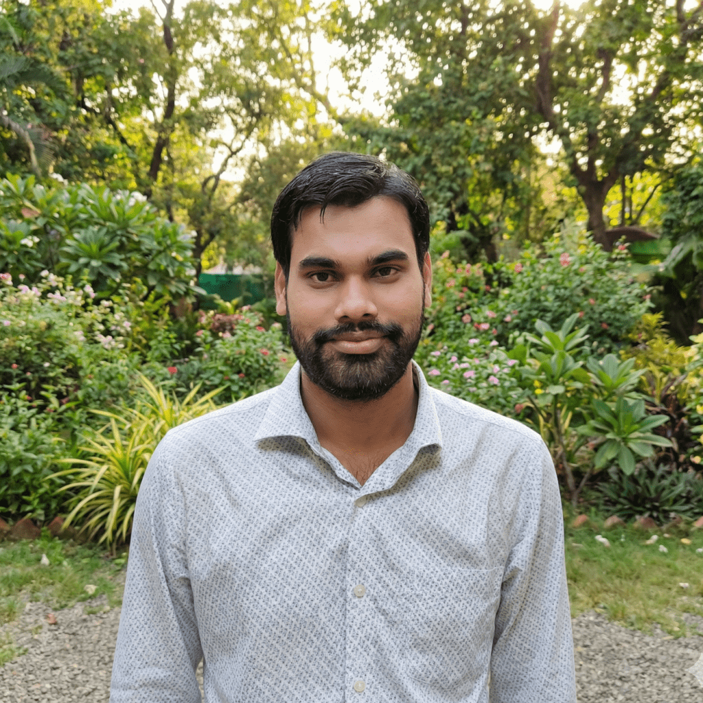 A young man with a beard wearing a patterned shirt standing in a garden with greenery and flowers.