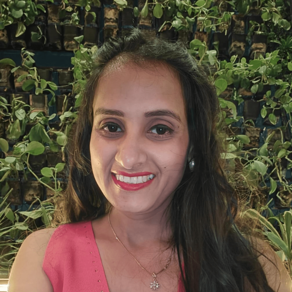A zoomed in shot of a smiling women, wearing a pink top, sitting in front of lush green garden with various plants and trees under natural light.
